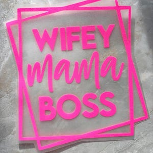 May include: A transparent sheet with the words "WIFEY Mama BOSS" in bright pink, set within a pink square frame. The text is in a bold, sans-serif font, with "Mama" in a cursive script. The background is a light gray, textured surface.