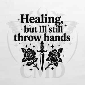 May include: Black and white graphic design with the text "Healing, but I'll still throw hands." The design includes a dagger, two roses, stars, and a bat-like shape.