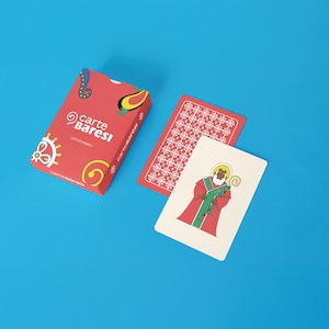 Può includere: Un mazzo di carte rosse "Carte Baresi" con un design decorativo. Vengono mostrate due carte, una con un motivo rosso e bianco e l'altra con un santo in abito rosso. La scatola e le carte sono su una superficie blu.