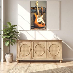 May include: An orange electric guitar with a white pickguard and a brown strap leans against a black amplifier. The guitar is the focal point of a framed artwork above a wooden cabinet with woven door panels. A potted plant and a round jute rug complete the scene.