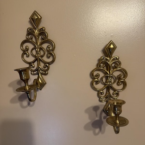 May include: A pair of polished brass wall sconces featuring an ornate, symmetrical scrollwork design with a diamond-shaped finial at the top and a circular candle cup holder extending from the base of each decorative metal wall-mounted fixture.