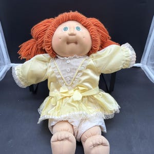 Könnte beinhalten: Eine Puppe mit leuchtend orangefarbenem Wollhaar in Zöpfen, grünen Augen und einem weichen Stoffgesicht. Die Puppe trägt ein hellgelbes Satinkleid mit weißem Spitzenkragen, drei kleinen weißen Knöpfen und einer passenden gelben Taillenschleife.