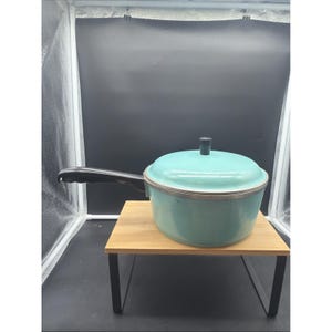 Puede incluir: Una olla redonda de color turquesa claro con tapa a juego y un mango largo de color negro. La olla presenta un borde de metal plateado en la parte superior y un pequeño pomo cilíndrico negro centrado en la tapa para su manipulación.