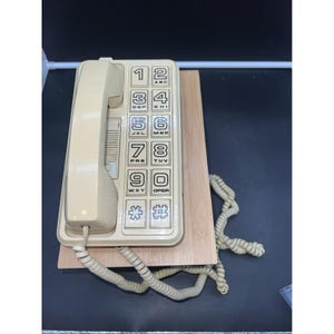 May include: A vintage beige wall phone with a matching handset. The phone has a rectangular keypad with numbered buttons and a floral and hash symbol. It is mounted on a light wood base with a coiled cord.