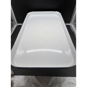 Puede incluir: Una bandeja rectangular de esmalte blanco con esquinas redondeadas y un borde azul oscuro. La superficie es lisa y brillante con un acabado reflectante. Los bordes muestran ligeros signos de desgaste y una pequeña decoloración.