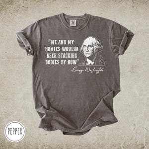 May include: A gray t-shirt with the quote "ME AND MY HOMIES WOULDA BEEN STACKING BODIES BY NOW" and a portrait of George Washington. The shirt has rolled-up sleeves and a crew neck.