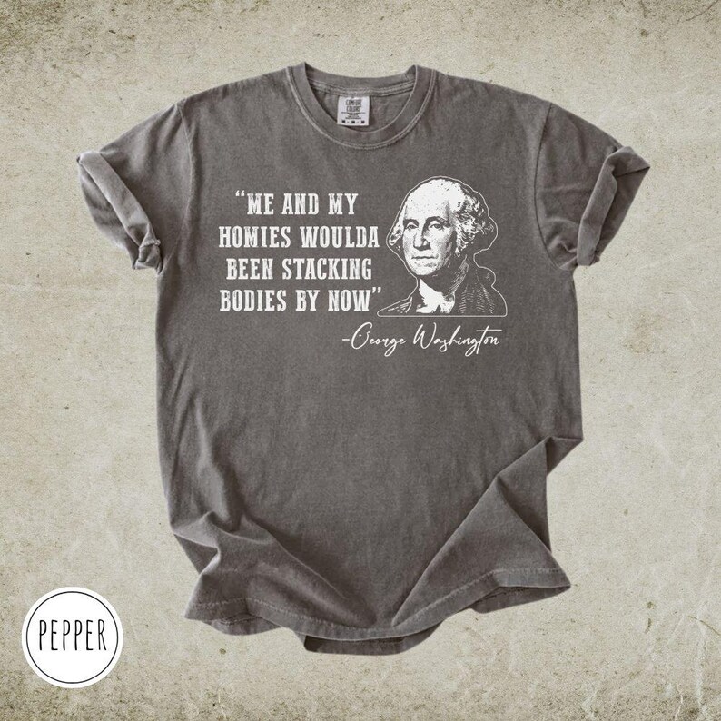 May include: A gray t-shirt with the quote "ME AND MY HOMIES WOULDA BEEN STACKING BODIES BY NOW" and a portrait of George Washington. The shirt has rolled-up sleeves and a crew neck.