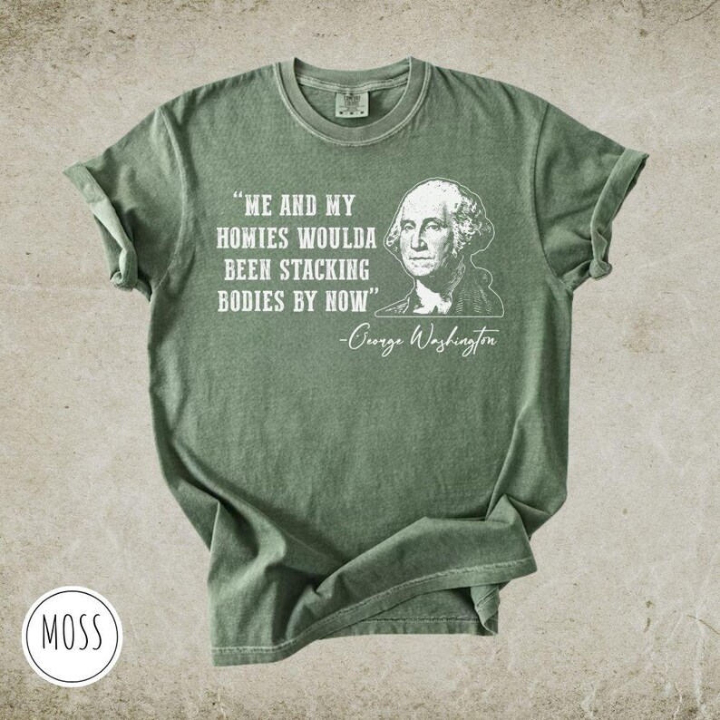 May include: Moss green t-shirt with rolled-up sleeves. The shirt features a white graphic of George Washington and the quote "ME AND MY HOMIES WOULDA BEEN STACKING BODIES BY NOW". The shirt has a round neckline and a relaxed fit.