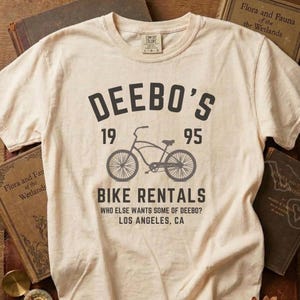 May include: A cream-coloured t-shirt featuring a bicycle graphic and the text "DEEBO'S BIKE RENTALS 1995 LOS ANGELES, CA". The shirt has a crew neck and short sleeves. Vintage books are in the background.
