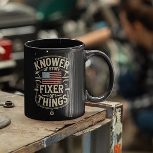 May include: Black ceramic mug with the text "Knower of Stuff, Fixer of Things" and an American flag design. The mug has a large handle and is sitting on a work surface. The background is blurred, showing a person working on a vehicle.