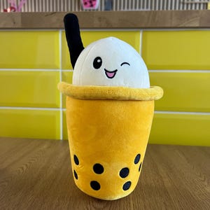 May include: A plush boba tea toy with a winking face and a black straw. The yellow cup has black bubble accents. A smaller boba tea plush is in the background.