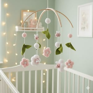 Op de afbeelding: Een baby mobile met een houten boog en hangende viltversieringen. De mobile heeft roze bloemen, witte pompons, groene bladeren en kleine bijtjes. Het hangt boven een wit babybedje.