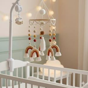 Könnte beinhalten: Ein Baby-Mobile mit Regenbogen- und Quasten-Dekorationen, die an einem Holzring hängen. Das Mobile wird von einem weißen Arm getragen und hängt über einem weißen Kinderbett. Die Regenbogen sind in Gelb-, Orange- und Cremetönen gehalten.