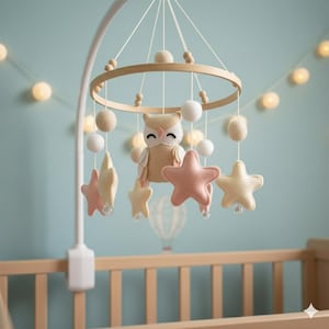 May include: A baby crib mobile featuring a wooden ring with hanging felt stars, spheres, and an owl. The stars are in shades of pink and yellow. The owl has a smiling face. The mobile is suspended above a wooden crib.