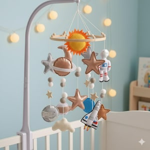 Op de afbeelding: Een baby mobile met vilt planeten, sterren, een zon, een astronaut en een raket. De mobile hangt aan een witte arm die aan een wieg is bevestigd. De planeten zijn grijs, bruin en blauw, met gouden en zilveren sterren.