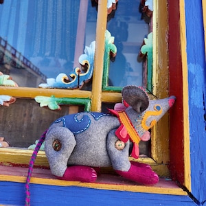 May include: A handcrafted gray felt mouse toy with blue and pink accents. The mouse has a long, striped tail and button details. It is posed on a colorful window frame with ornate carvings.