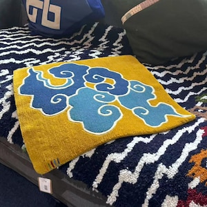 Pode incluir: Uma capa de almofada quadrada com base amarelo-mostarda e um design central bordado de nuvens estilizadas em tons de azul-escuro e azul-claro. A textura parece ser tufada ou tecida, com uma pequena etiqueta multicolorida visível no canto.