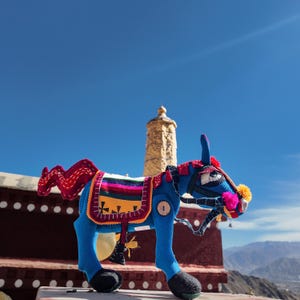 Könnte beinhalten: Eine blaue Eselfigur aus Stoff mit einem roten, gewellten Schwanz, einer bunten, gestreiften Satteldecke mit geometrischen Mustern und einem Zaumzeug, das mit rosa und gelben Pompons verziert ist. Der Esel hat schwarze Hufe und einen runden Holzknopf.
