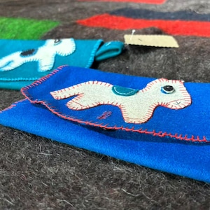 Könnte beinhalten: Zwei rechteckige Filztaschen in Blautönen, jeweils mit einer aufgenähten Applikation eines vierbeinigen Tieres. Die vordere Tasche ist königsblau mit roten Deckenstichen und einem cremefarbenen Tier mit türkisfarbenem Sattel.