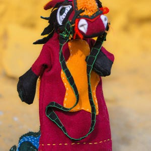 May include: A handmade felt dragon puppet with a red body, black arms, and a colorful head. The dragon has a bright orange chest, a green and black harness, and a blue and green tail. The puppet is mounted on a black stand.