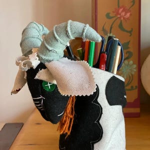 May include: A cylindrical fabric pencil holder featuring a black and white goat head design. The goat has light green horns, a green button eye with a white cross, white fabric ears, and an orange yarn beard. It holds various pens and pencils.