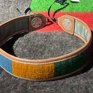 May include: A camera strap featuring a multi-colored felt exterior with blue, mustard yellow, and green rectangular panels. The strap is bordered by tan leather trim with white stitching and includes two tan leather end tabs with a circular white logo.