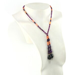 May include: A beaded necklace displayed on a white mannequin. The necklace features a combination of purple, orange, and black beads. The necklace has a Y-shaped design with two dangling strands ending in dark, faceted beads.