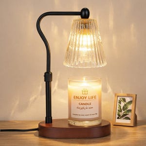 Può includere: Una lampada scaldacandele in metallo nero con paralume in vetro scanalato e base in legno scuro. Un barattolo di candela in vetro trasparente con l'etichetta ENJOY LIFE CANDLE poggia sulla base. La lampada emette una luce calda che illumina la candela.