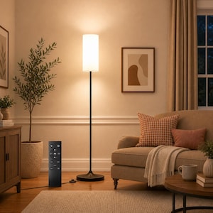 May include: A tall floor lamp with a slender black metal pole and a cylindrical white fabric shade that emits a warm glow. The lamp features a circular black base and is accompanied by a black rectangular remote control with various buttons.