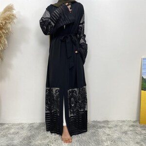 May include: A black open-front abaya with intricate lace and sheer detailing on the sleeves and hem. The abaya features a matching belt and is worn over white pants. The sleeves have a pleated design, and the garment is floor-length.