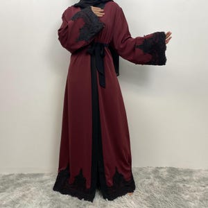 以下が含まれることがあります： 袖と裾に黒いレースのディテールが施された、バーガンディ色のアバヤ。アバヤは黒いベルトと前面に黒いトリムが特徴です。袖は長くフレアで、衣服は床の長さです。