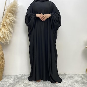 以下が含まれることがあります： 床まで届く長さの黒いアバヤで、ゆったりとした流れるようなシルエットが特徴です。袖は長く幅広で、手首部分はゴム入りのカフスで絞られています。ハイネックのラウンドカラーで、不透明な無地の生地が裾まで垂れ下がっています。