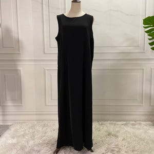 Può includere: Abito lungo nero senza maniche. L'abito ha uno scollo tondo semplice e una silhouette ampia e fluida. Viene presentato su un manichino in uno studio con un tappeto bianco e una parete.