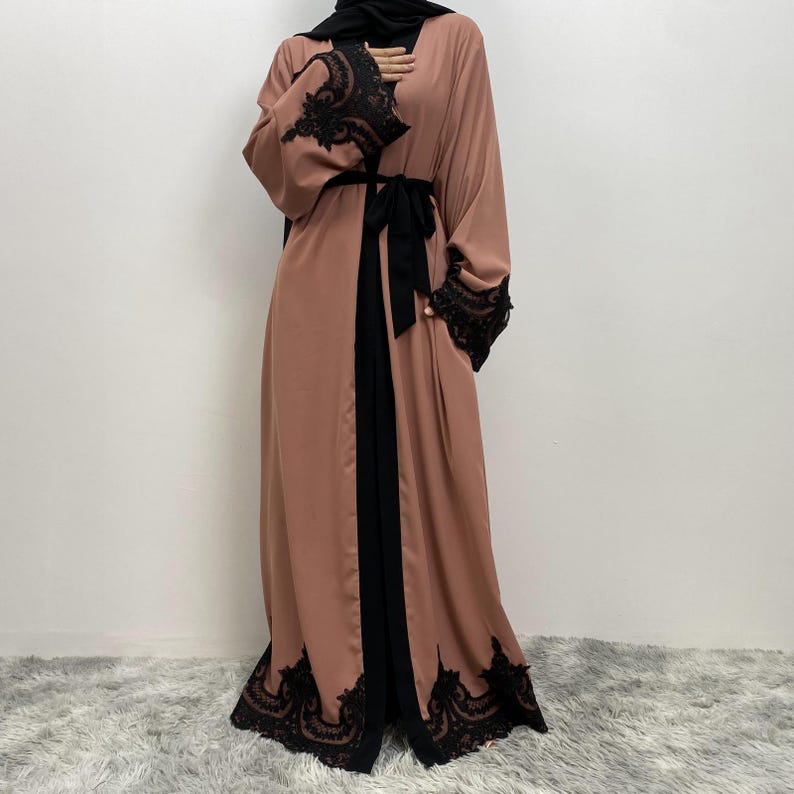 以下が含まれることがあります： 袖と裾に黒いレースのトリムが付いた、長くて流れるようなピーチ色のアバヤ。アバヤは前面に黒いパネルと、お揃いの黒いベルトが特徴です。長くてゆったりとした袖で、全体的に控えめでエレガントなスタイルです。