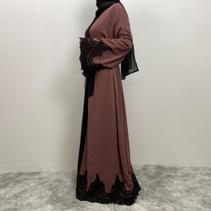 以下が含まれることがあります： 袖と裾に黒いレースのトリムが付いた、長く流れるようなモーヴ色の abaya。 abaya は黒いベルトが特徴で、黒いヒジャーブと組み合わせています。 袖には複雑なレースのディテールがあります。