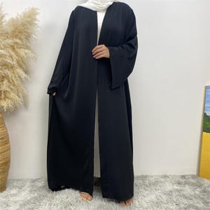 May include: A long, solid black open-front abaya featuring wide, flowing sleeves and a smooth, matte fabric finish. The garment is worn over a white base layer and is paired with a white head covering, creating a simple, layered silhouette.