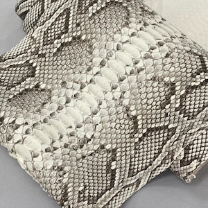 Può includere: Primo piano di un cuscino decorativo con motivo a pelle di serpente. Il cuscino presenta un motivo ripetuto di squame bianche, grigie e marroni. La trama del cuscino è visibile e il design è simmetrico.