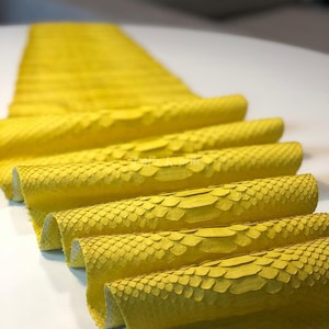 May include: Close-up of several rolls of bright yellow leather. The leather has a textured, scale-like pattern, and the edges of the rolls are visible. The rolls are arranged in a row, with some overlapping. The background is a blurred white surface.