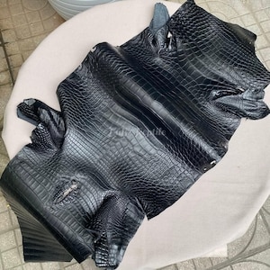 May include: A large piece of black crocodile leather with a textured, scaly pattern. The leather is folded and draped, revealing the natural shape and texture of the material. The image showcases the luxurious quality of the exotic leather.