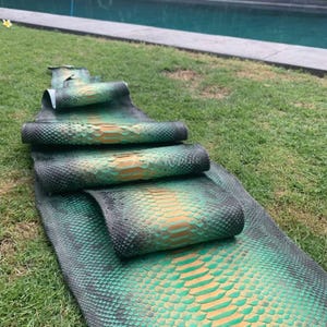 May include: Several pieces of dyed snakeskin in shades of green, black, and orange are arranged on green grass. The snakeskin is rolled up in sections, revealing the textured scales and color variations. The snakeskin is likely for crafting.