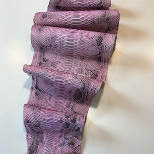 Può includere: Un pezzo arrotolato di pelle di serpente rosa e grigia. La pelle di serpente ha un motivo a scaglie ed è un mix di colori rosa, lavanda e grigio. La pelle è ripiegata su se stessa, mostrando la consistenza e il motivo.