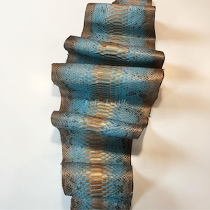 May include: A length of reptile skin in teal, brown, and gold hues. The scales are clearly visible, and the texture looks rough. The text "Felix Reptile" is printed on the skin. This material is likely used for crafting purposes.