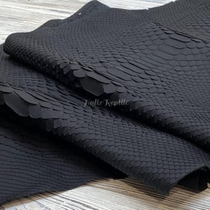 Puede incluir: Primer plano de cuero de reptil negro con un patrón de escamas. El cuero está doblado, revelando la textura y el brillo. El texto "Hafiz Reptile" es visible en la esquina inferior derecha.