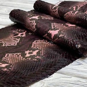 May include: Close-up of a rolled-up piece of dark burgundy leather with a snakeskin pattern. The pattern features pink and black scales. The leather appears to be high-quality and suitable for various crafting projects.