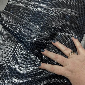 May include: A close-up of dark blue snakeskin leather with a scale-like pattern and a glossy sheen. The leather is folded, revealing its underside. A hand with a ring is touching the leather. The texture is clearly visible.