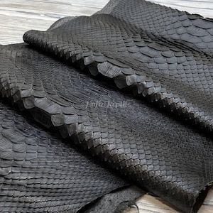 Op de afbeelding: Close-up van verschillende vellen zwart pythonleer. Het leer heeft een getextureerd, geschubd patroon en een matte afwerking. De vellen zijn gelaagd en tonen de natuurlijke plooien en variaties in het materiaal. De algehele uitstraling is donker en luxueus.