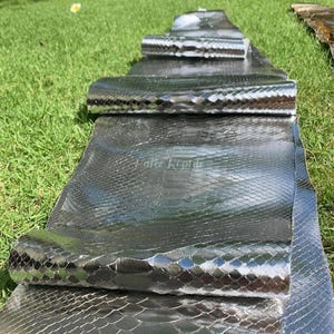 Peut inclure: Plusieurs rouleaux de cuir de peau de serpent gris foncé brillant sont disposés sur de l'herbe verte. Le cuir présente un motif d'écailles et une surface réfléchissante. Le texte "Fatte Reptile" est visible sur le cuir.