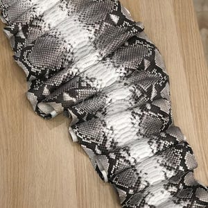 Può includere: Un pezzo piegato di pelle di serpente nei toni del grigio, nero e bianco. Il motivo ricorda le squame di un pitone. La pelle è appoggiata su una superficie di legno chiaro.