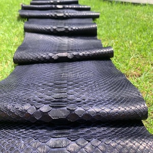 May include: Close-up of several rolls of dark blue snakeskin leather. The leather has a textured, scale-like pattern and is arranged on a bed of green grass. The image is well-lit, highlighting the details of the material.
