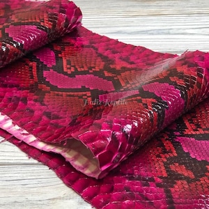 Peut inclure: Gros plan sur un morceau de cuir exotique texturé. Le matériau présente un motif d'écailles rouges, roses et noires, avec une finition brillante. Le texte "Taliz Reptile" est visible sur le cuir.